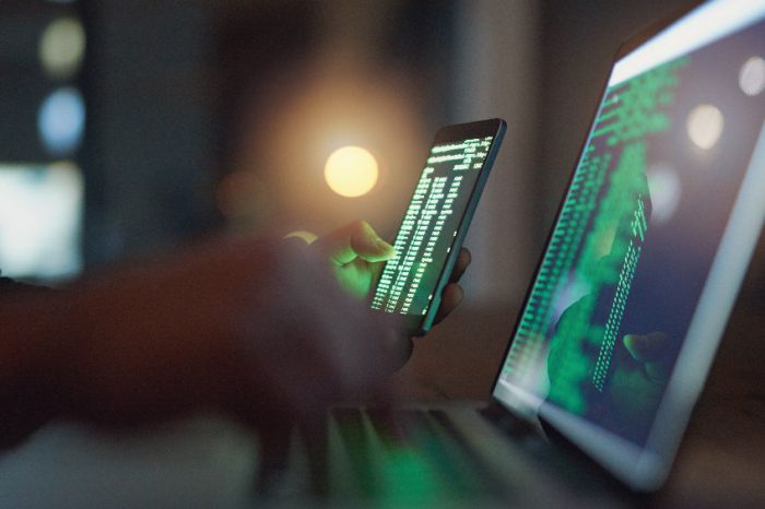 A close-up of a person’s hands using a smartphone and a laptop with screens displaying lines of green code