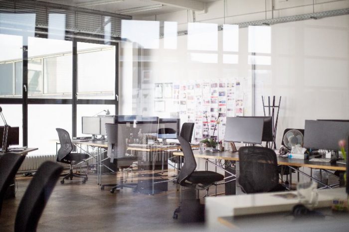 A modern office space with multiple desks and computers, ergonomic chairs and a whiteboard, viewed through a glass partition