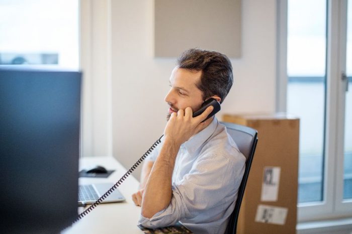 An individual sitting at a desk in an office environment, holding a telephone to their ear, with a computer monitor on the desk