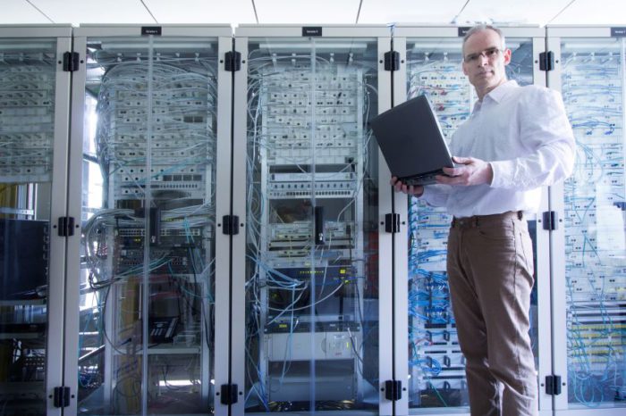 A person standing in a data center or server room, holding an open laptop and working on it, with multiple server racks and various cables and equipment in the background