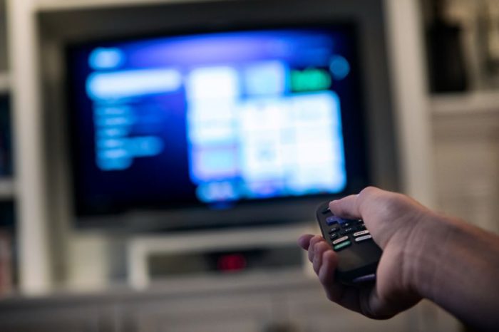 A person’s hand holding a television remote control, pointing it towards a TV screen displaying a menu interface