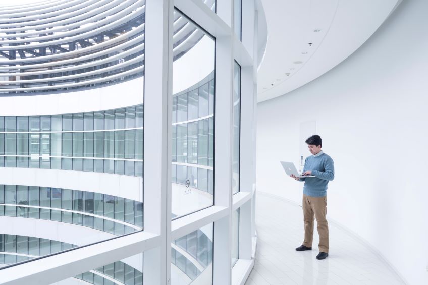 An individual stands in a modern building, holding and looking at an open laptop