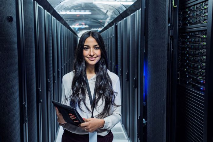 An individual stands in a data center aisle between server racks, holding a tablet