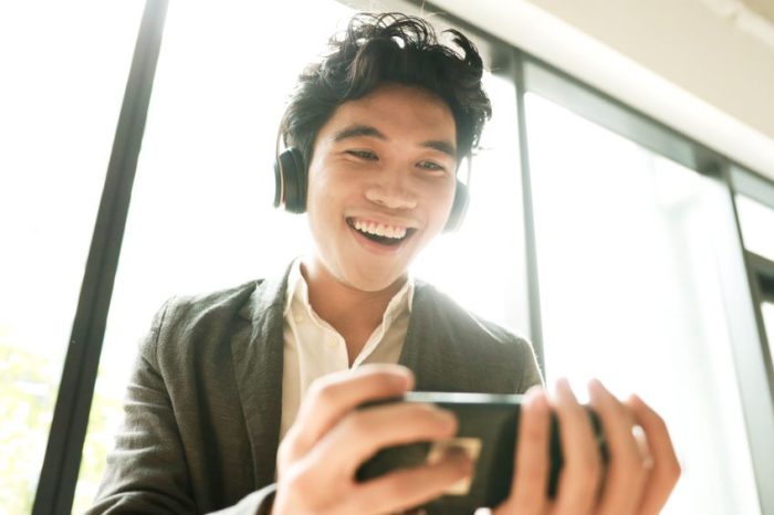 An individual wearing a dark-colored blazer and a white shirt, with headphones on, holding and looking at a smartphone