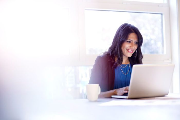 An individual sits at a table using a laptop with a cup beside it, with a window and bright light in the background