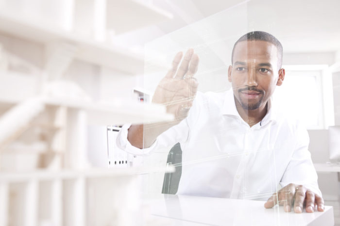 An individual sits at a desk with their right hand raised in a stopping gesture, interacting with a transparent interface full of geometric shapes and lines