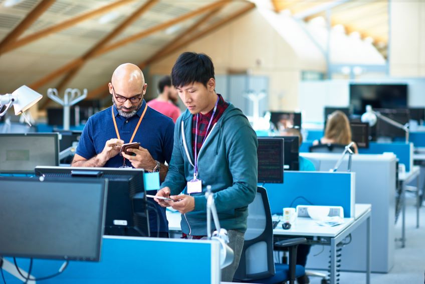 An indoor office environment with multiple workstations, where two individuals are standing in the foreground and looking at their phones