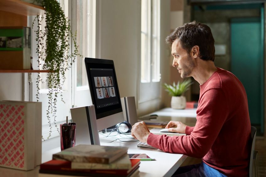 An individual sits at a desk in front of a computer displaying a video conference call, with various items like books, a notebook, a cup and glasses on the desk