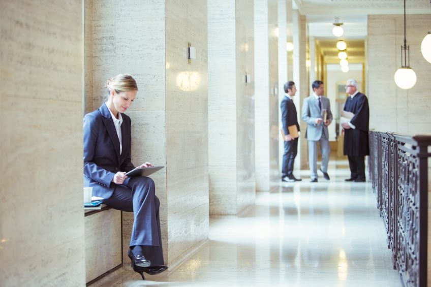 An individual sits on a ledge in a hallway, holding a tablet, while three others stand and converse in the background of a formal building with classical architecture