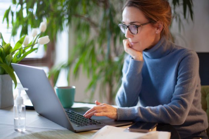 An individual sits at a table using a laptop, with a cup, a bottle of hand sanitizer and some papers beside the laptop