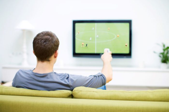 An individual sits on a green sofa, holding a remote control and facing a television screen displaying a soccer match