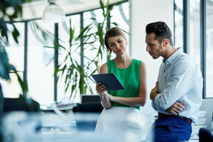 Two individuals in a professional setting, while one holds a tablet and the other observes what's on it