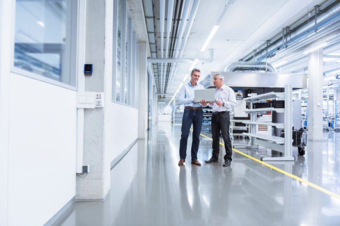 Two individuals in an industrial setting, talking to one another as one of them holds a laptop