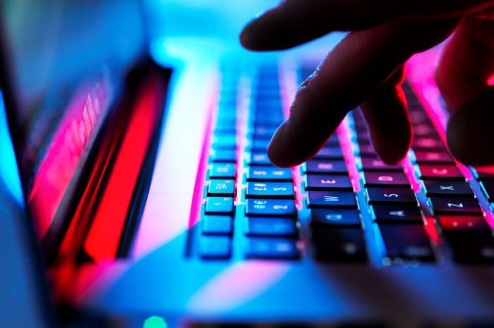 A close-up of a person’s hand typing on a laptop keyboard, which is backlit with blue and red colors