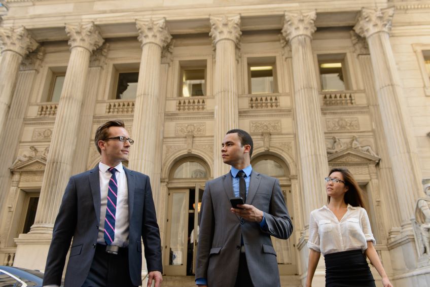 Three individuals in professional attire are walking outside a building with classical architecture featuring columns and steps