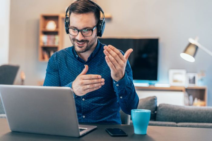 An individual sitting at a table with a laptop open in front of them, wearing headphones and gesturing with their right hand