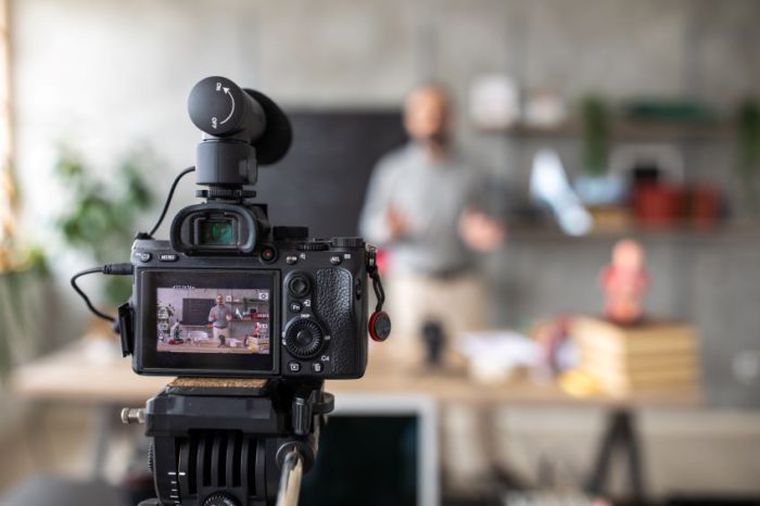 A digital camera on a tripod, focused on a person giving a presentation in front of a kitchen counter