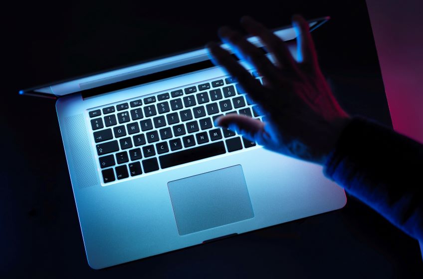 A laptop with an illuminated keyboard is shown in a low-light environment while a hand hovers above the keyboard