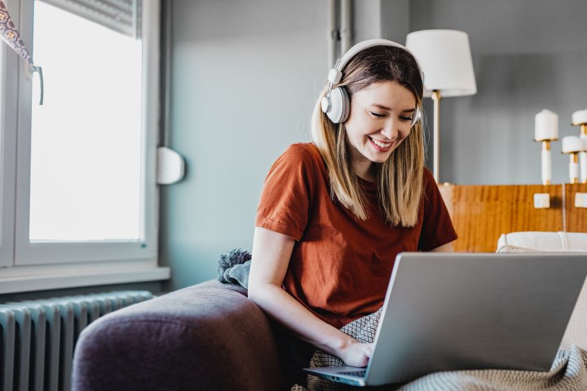 A person is seated on a purple couch while using a laptop and headphones