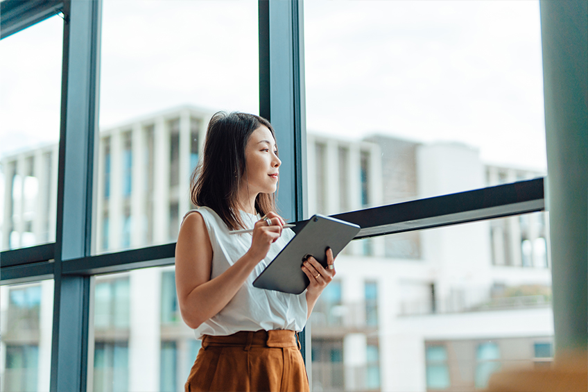 An office professional looks out a window thinking about work while holding a tablet and stylus.