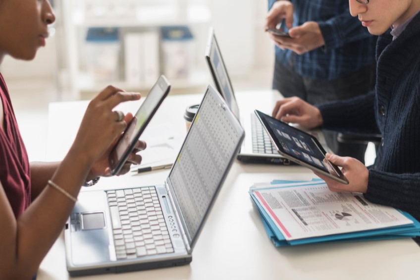 Three people using smart devices working together on spreadsheets and pie charts.