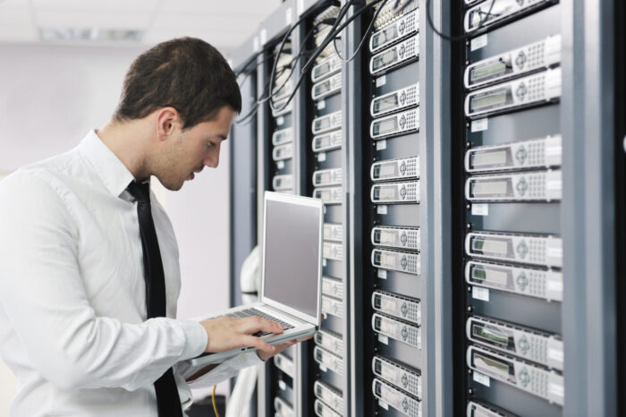 IT specialist standing in front of a row of servers in a data center holding an open laptop