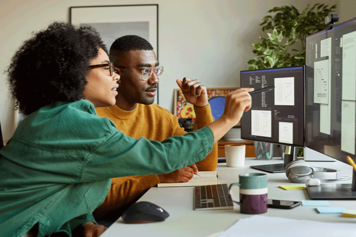 Two people sitting at a desk looking at information on computer monitors as one of them points the screen