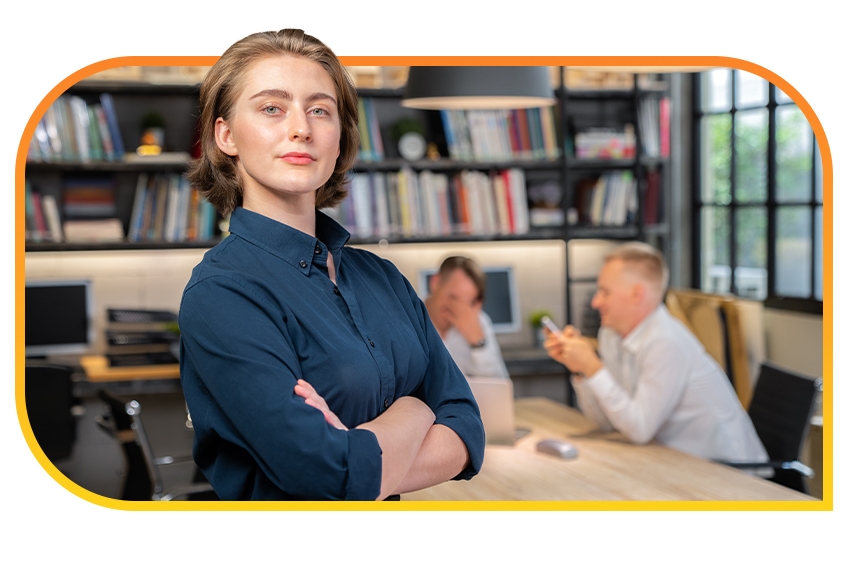 A professional woman with crossed arms stands confidently in an office space with bookshelves and two people sitting at a shared desk in the background