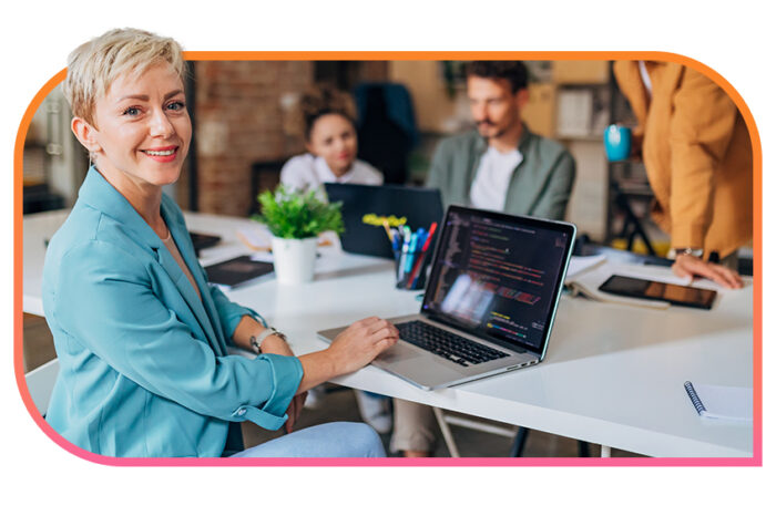 A woman in a modern office space sits at a shared desk and smiles while using a laptop