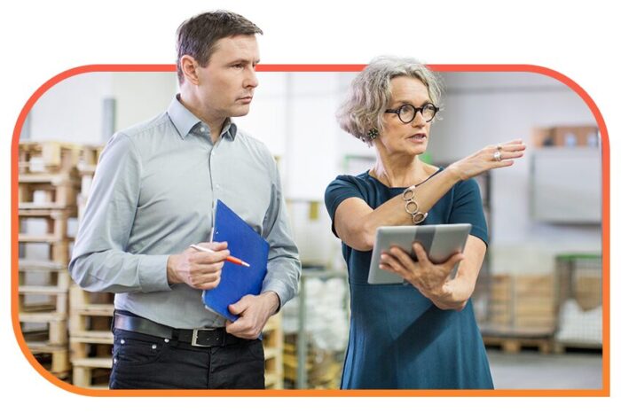 Two people standing together in a warehouse holding tablets and notebooks while talking.