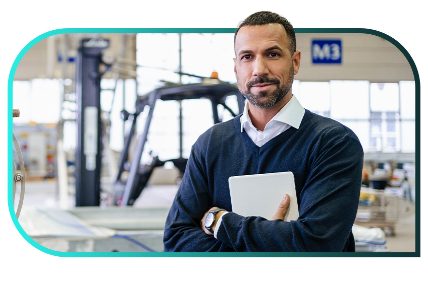 A man in casual business attire holds a tablet in crossed arms in a brightly lit factory space