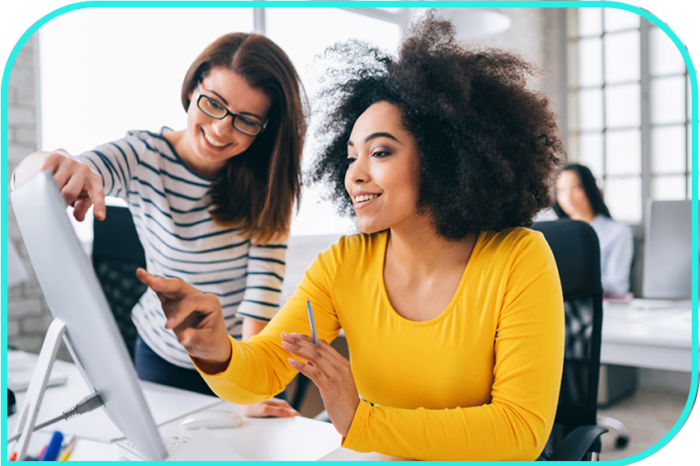 Two women in casual business attire look at a computer and talk in an office