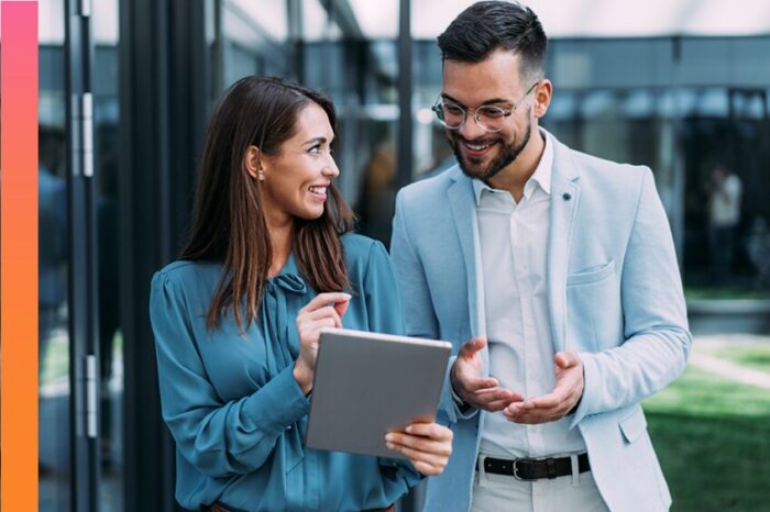 A man and a woman in casual business attire smile as they talk and study a tablet outside an office building