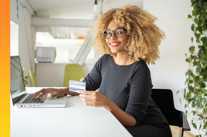 Woman sitting at desk with laptop open completing an online transaction with credit card in one hand while typing with the other.