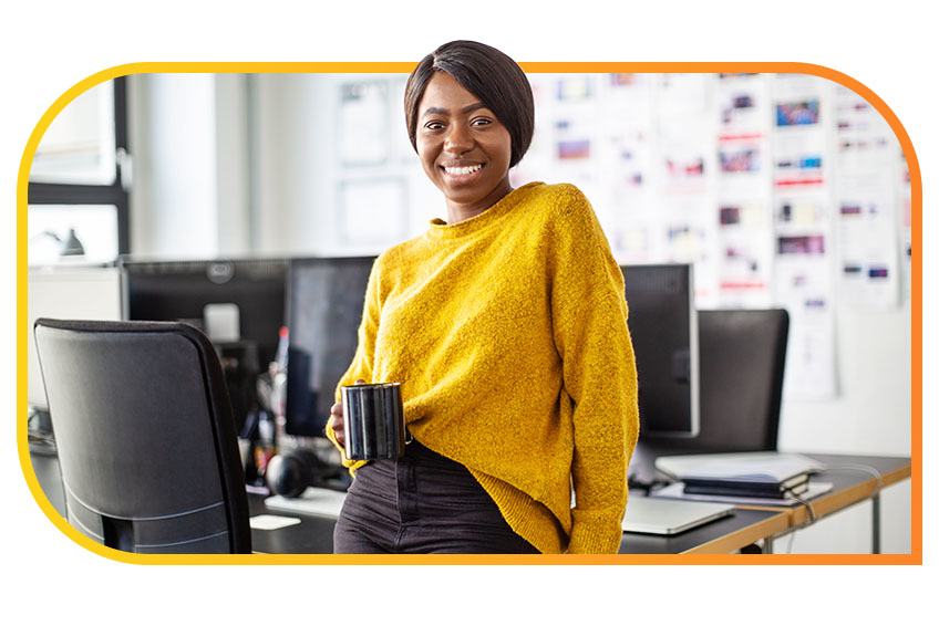 A woman holding a mug leans against a table in an office space and smiles