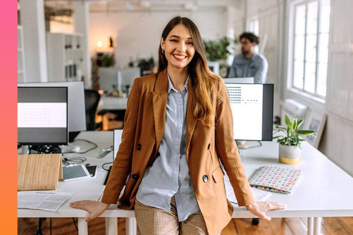 A woman in casual business attire smiles and leans against a table with computers in a modern office space.