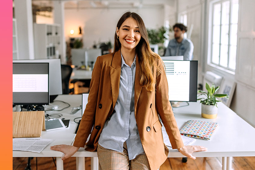 A woman in casual business attire smiles and leans against a table with computers in a modern office space.