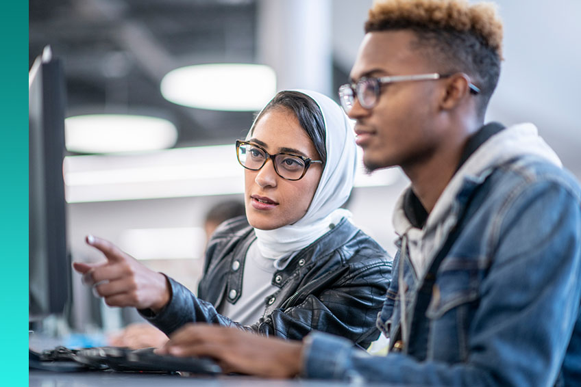 Two individuals engaged in a conversation as one gestures towards something on a computer monitor