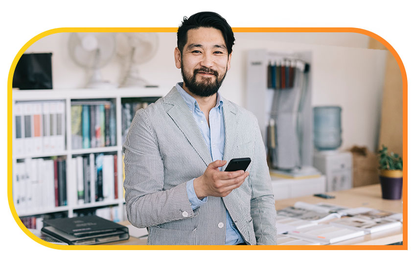 Man leaning against a desk with phone in hand smiling and looking directly into camera very pleased with what he just accomplished.