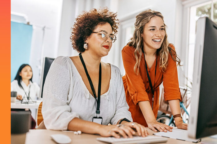 Two female colleagues reviewing information from a large monitor at an office desk.