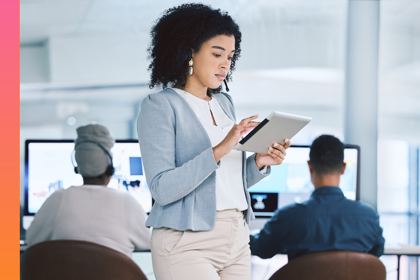 Person holding a tablet and standing in front of two people sitting at computer desks.