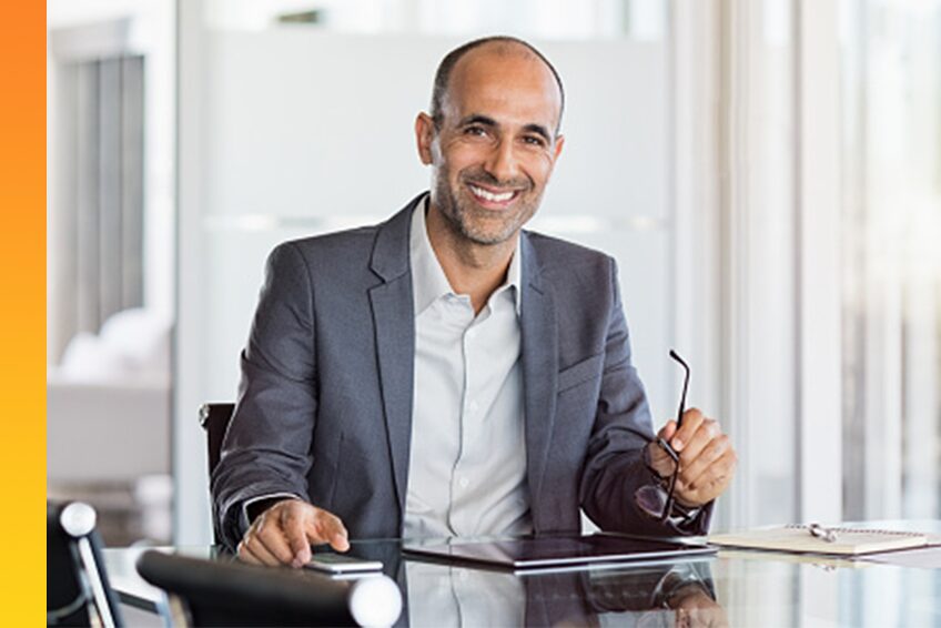 Man sitting at glass office desk with spectacles in hand smiling after reviewing a favorable report on computer tablet.