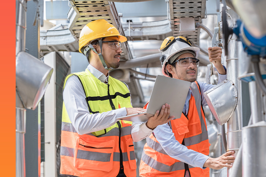 Two individuals in high-visibility vests and hard hats, one holding a laptop, inspecting water system equipment.