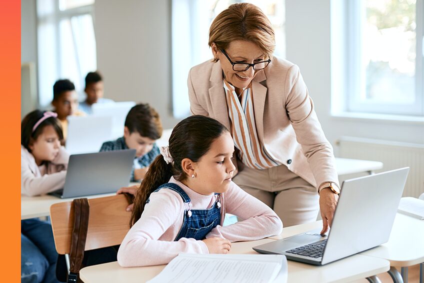 A teacher smiles and talks to a student who is sitting at a desk with a laptop